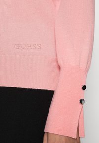 Manche de pull en tricot rose avec deux boutons noirs sur le poignet et le logo "GUESS" brodé en petites perles claires près de l'ourlet.
