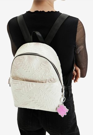 Witte, gestructureerde rugzak met zigzagpatroon en zilverkleurige ritssluiting. Voorzien van een roze sleutelhangeraccent bevestigd aan de rits. Zwarte bandjes.
