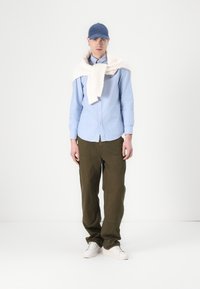 Lichtblauwe button-up overhemd, olijfgroene broek, witte kabelgebreide trui over de schouders gedrapeerd, blauwe pet, witte sneakers. Minimalistische achtergrond.