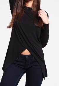 Maglietta nera a maniche lunghe con design incrociato, caratterizzata da un orlo obliquo. Indossata con jeans blu scuro che mostrano una leggera lucentezza sul tessuto.