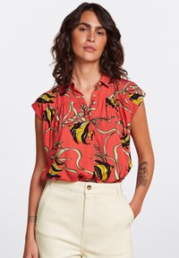 Camisa roja de botones con un patrón floral y de peces en negro y amarillo, con mangas cortas y un corte holgado, combinada con pantalones de color claro.