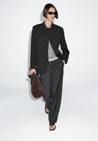 Blazer oversize a righe nere, top grigio a strati, pantaloni scuri, borsa in suede marrone, sandali neri minimalisti, sfondo neutro.
