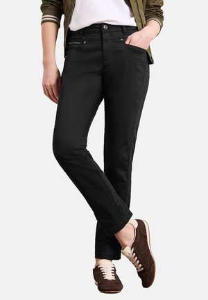 Person iført sorte slim-fit bukser, hvid skjorte, olivengrøn jakke og brune sneakers, stående med den ene hånd i lommen og benene krydsede.