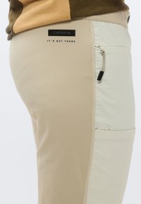 Beige bukser med en struktureret overflade. Har en sidelomme med lynlås og et kontrasterende lys beige panel. Logo-tag over lommen.