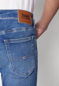 Modré džíny z denimové tkaniny s hnědým koženým štítkem označeným "TOMMY JEANS" v pase. Obsahují zadní kapsu s malým logem.
