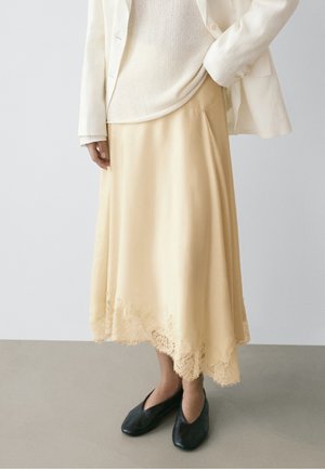 Femme portant une jupe midi crème bordée de dentelle, un haut en maille blanc, un blazer blanc et des chaussures plates noires, debout sur un sol uni.