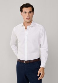 Camisa blanca de cuello con tapeta de botones, mangas largas, textura suave. Combinada con pantalones marinos y un cinturón de cuero marrón.