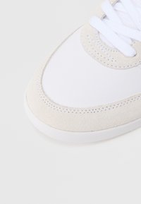 Detailní záběr přední části bílého tenisky s béžovými semišovými detaily a bílými tkaničkami na jednobílém pozadí.