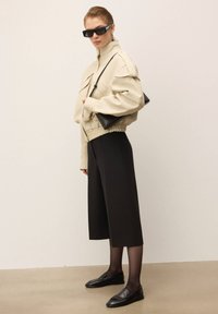 Veste cropped beige avec un ourlet élastique, pantalon noir à jambes larges et mocassins noirs. L'accessoire comprend un petit sac à main noir.