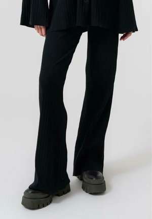 Pantalones acampanados de canalé negros, hechos de material suave, con una cintura alta y combinados con zapatos gruesos de suela oscura.