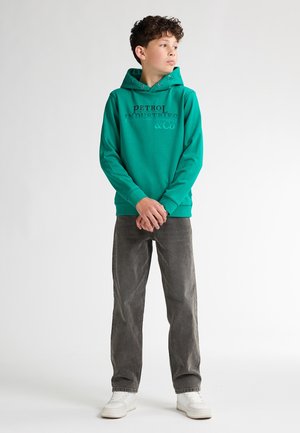 Groene hoodie met zwarte tekst, gecombineerd met grijze jeans en witte sneakers. De hoodie heeft een kangoeroe-pocket en een trekkoordkap. 
