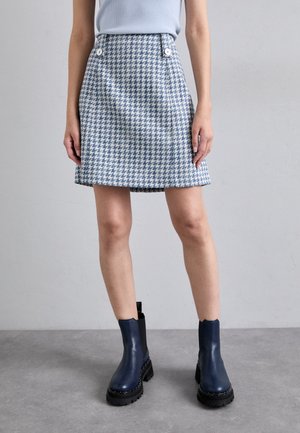 Sininen ja valkoinen houndstooth-kuvioinen hame, jossa on kaksi etunappia, liehuva muoto ja sileä rakenne, yhdistettynä laivastonsinisiin nilkkureihin.