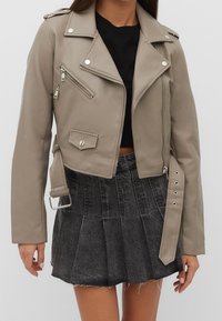 Beige faux leather biker jacket with silver zippers and studs; features a cropped design, asymmetrical front, and zippered pockets.