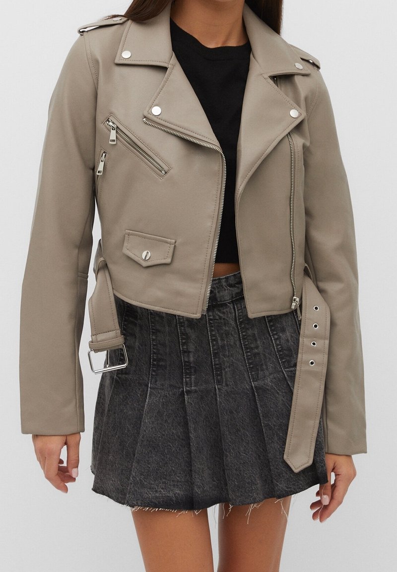 Beige faux leather biker jacket with silver zippers and studs; features a cropped design, asymmetrical front, and zippered pockets.