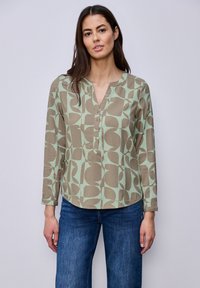 Femme portant un chemisier à manches longues vert et beige à motifs et un jean bleu, debout contre un mur clair uni.