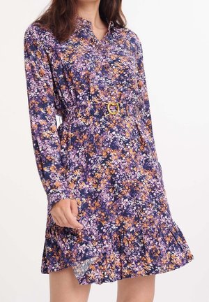 Donna che indossa un vestito a maniche lunghe, lungo fino al ginocchio, con motivo floreale viola, arancione e bianco e fibbia della cintura circolare dorata.