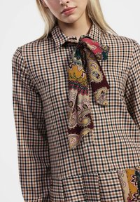 Abito a scacchi marrone e beige con un fiocco al colletto, caratterizzato da un motivo paisley in tonalità di bordeaux, giallo e verde.