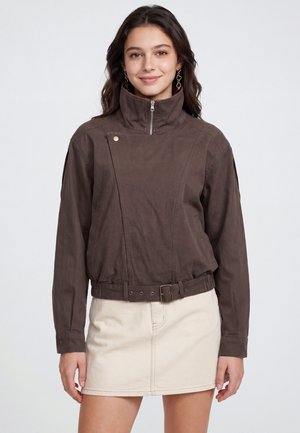 Giovane donna che indossa una giacca marrone con zip e cintura e una gonna color crema, sorride con capelli lunghi e scuri e orecchini a catena.