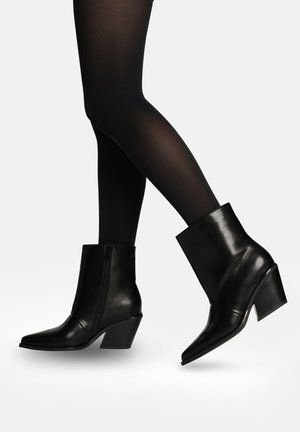 Bottes bottines en cuir noir avec bouts pointus et talons épais portées avec des collants noirs transparents sur des jambes croisées.