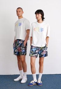 Hvide kortermede t-shirts med multicolor "adidas" logo, parret med mønstrede shorts i mørke nuancer med grafiske designs, vist på to modeller.