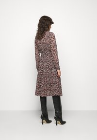 Mujer con cabello rizado vestida con un vestido floral de manga larga y botas de tacón negras por la rodilla, de espaldas contra un fondo liso.