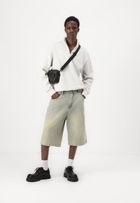 Sweatshirt cu fermoar gri deschis, shorts din denim estompați și pantofi negri masivi cu șireturi. Modelul poartă de asemenea o geantă mică de umăr neagră.