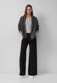 Vrouw die met handen in de zakken staat, gekleed in een grijze gestructureerde cardigan, zilveren tanktop, wijde zwarte broek en witte sneakers voor een grijze achtergrond.
