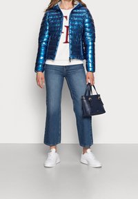 Vrouw in een glanzend blauwe donsjas, witte trui met rode letters, korte blauwe spijkerbroek, witte sneakers, houdend een marineblauwe handtas aan de zijkant.