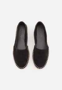 Scarpe slip-on in suede nero con cuciture beige lungo la suola, dotate di un interno liscio e di un design piatto per comfort e praticità.