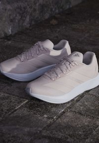 Baskets de sport rose clair avec une tige texturée, trois rayures sur le côté et une semelle blanche. Présente un design à lacets et un collier rembourré.
