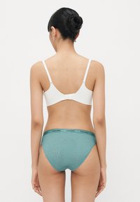 Bela braletka z nastavljivimi naramnicami in sponko na hrbtu, v kombinaciji z teal čipkastimi hlačkami z črnim pasom Calvin Klein.