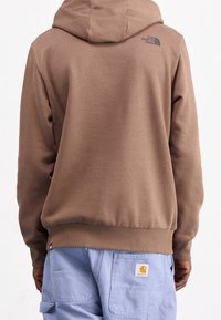 Osoba v hnědé mikině The North Face a světlých modrých kalhotách Carhartt, zobrazená zezadu na bílém pozadí.