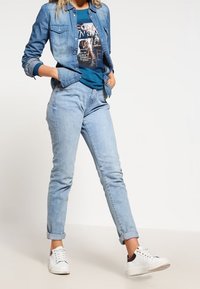 Ljusblå denimskjorta kombinerad med en teal grafik-t-shirt, ljusblå jeans och vita sneakers med bruna accenter. Avslappnad passform och uppvikta ärmar.