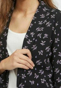 Schwarzer Blazer mit floralem Muster in Lavendel und Weiß. Verfügt über einen Reverskragen und einen taillierten Schnitt, getragen über einem weißen Oberteil.
