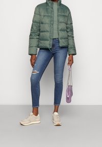 Giacca a vento verde con colletto alto, tessuto testurizzato e zip frontale, indossata sopra un maglione verde. Jeans skinny blu, sneakers chiare e una borsa viola.