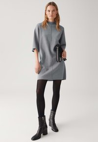 Frau in einem grauen knielangen Kleid mit halben Ärmeln, schwarzen Strumpfhosen, schwarzen Knöchelstiefeln, die eine kleine schwarze Tasche mit gestreiftem Tragegurt hält.