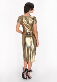 Metallisch gouden jurk met korte mouwen, aansluitende silhouet en een jurk tot op de knie; gecombineerd met zwarte hoge hakken met verenaccenten.
