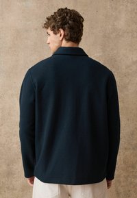 Ciemnoniebieska wełniana kurtka z kołnierzem, dopasowany krój i gładka faktura. Widok z tyłu ujawnia prosty design bez widocznych elementów metalowych czy ozdób.