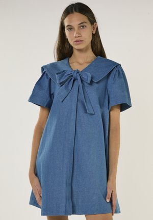 Jeune femme portant une robe en jean bleue avec des manches courtes bouffantes et un grand col marin noué en nœud, debout devant un fond uni.