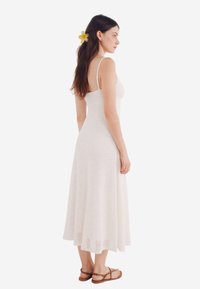 Robe blanche sans manches avec un motif texturé et une jupe évasée, dotée de fines bretelles. Le mannequin porte des sandales marron et a une fleur dans les cheveux.