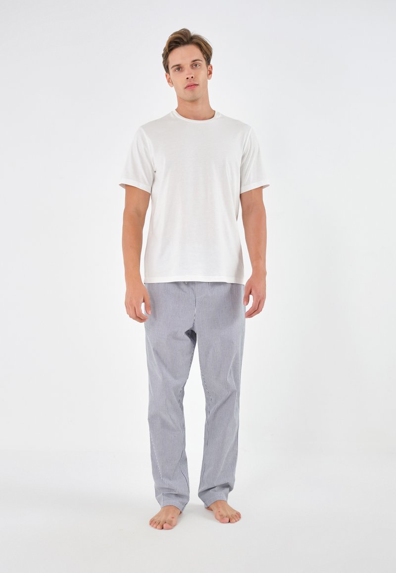 Tricou alb din bumbac și pantaloni de pijama gri cu dungi. Pantalonii au o croială lejeră și dungi verticale. Modelul stă desculț în fața unui fundal simplu.