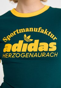 Green t-shirt with a yellow ribbed collar featuring bold printed text: "Sportmanufaktur adidas HERZOGENAUCH" and an Adidas logo.