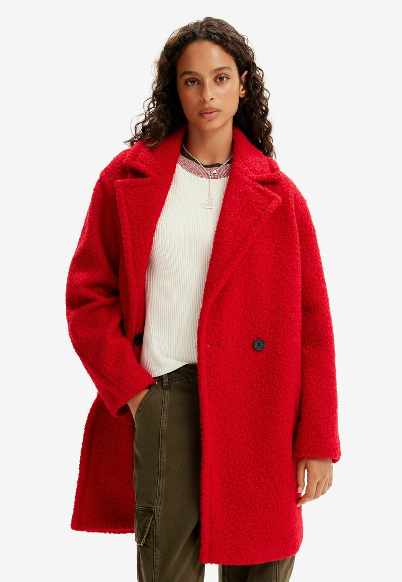 Cappotto oversized rosso in misto lana con finitura testurizzata, grandi risvolti e bottoni neri a contrasto; indossato sopra un top bianco a costine.