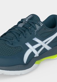 Σκούρο πράσινο-μπλε αθλητικό παπούτσι Asics με λευκά λογότυπα, κορδόνια, επάνω μέρος από δίχτυ και νεον κίτρινο λεπτομέρεια σε γκρι και λευκή σόλα.