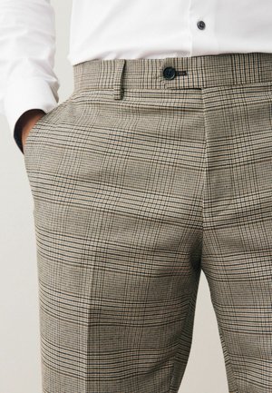 Muž v béžových a černých kostkovaných kalhotách s černým knoflíkem, rukama v kapsách, a bílou košilí s dlouhým rukávem zastrčenou dovnitř.