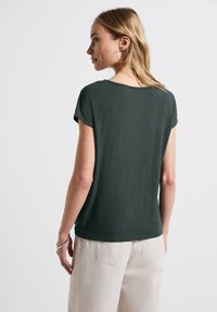 T-shirt a maniche corte verde scuro con una vestibilità rilassata e una texture morbida, abbinato a pantaloni beige chiaro. Design minimalista, senza motivi visibili.