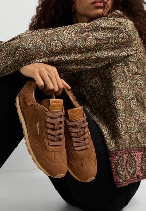 Persoon die een top met lange mouwen en patroon draagt, houdt een paar bruine suède sneakers vast met gumzolen en bruine veters.