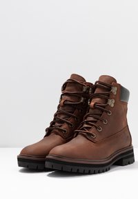 Botas de senderismo de cuero marrón con ojales metálicos, cordones negros, un cuello acolchado y una suela negra resistente con un patrón de textura.