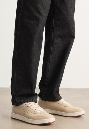 Zapatillas beige con parte superior de ante texturizado, media suela blanca y suela de goma marrón claro, combinadas con vaqueros de mezclilla negros con costuras sutiles.