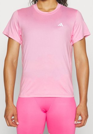 Osoba ubrana w jasnoróżową koszulkę Adidas z krótkim rękawem i jaskrawo różowe sportowe krótkie spodenki, pokazana od szyi do połowy uda, stoi z rozluźnionymi ramionami.